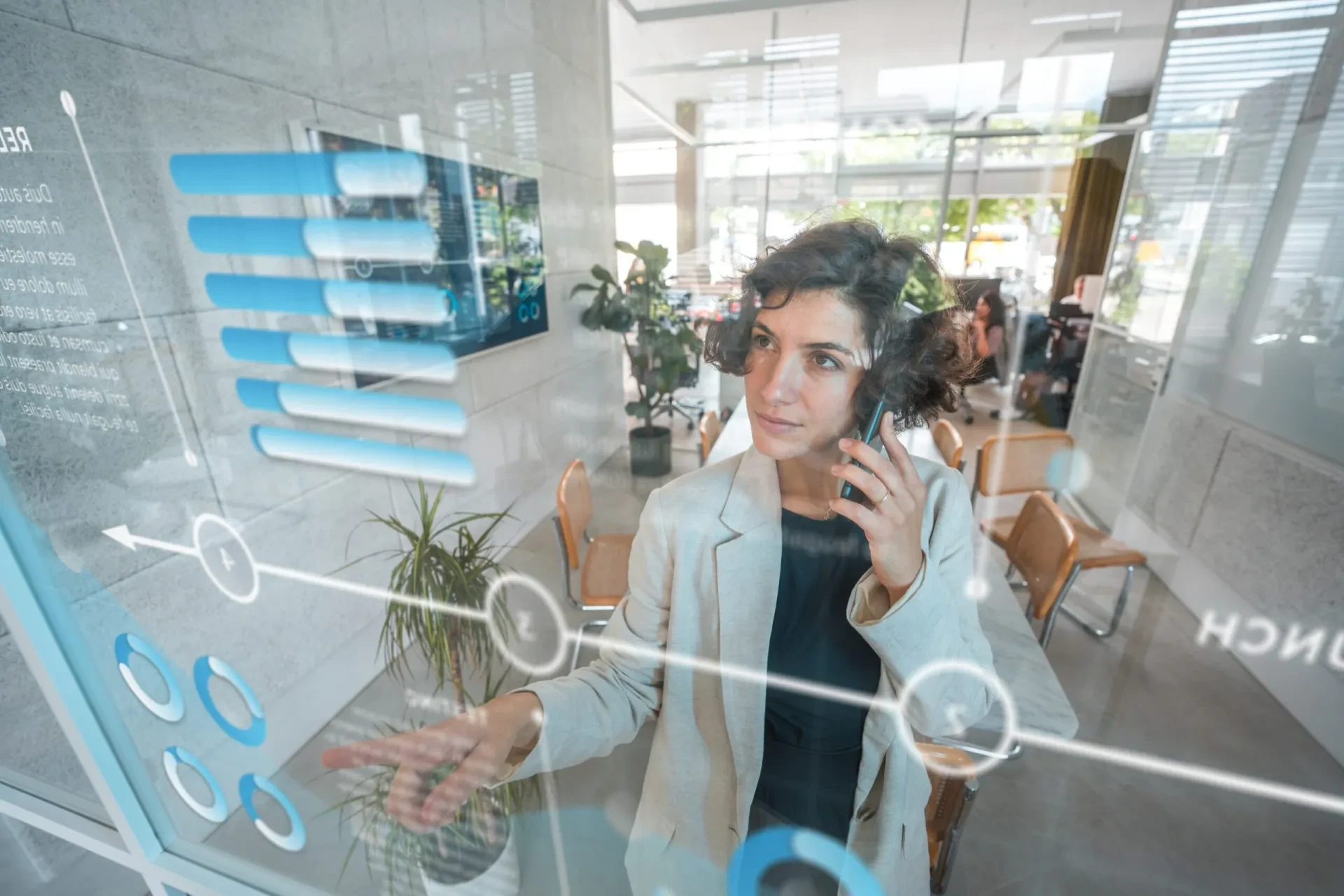Frau telefoniert und zeigt auf digitale Diagramme auf einer Glasscheibe im Büro