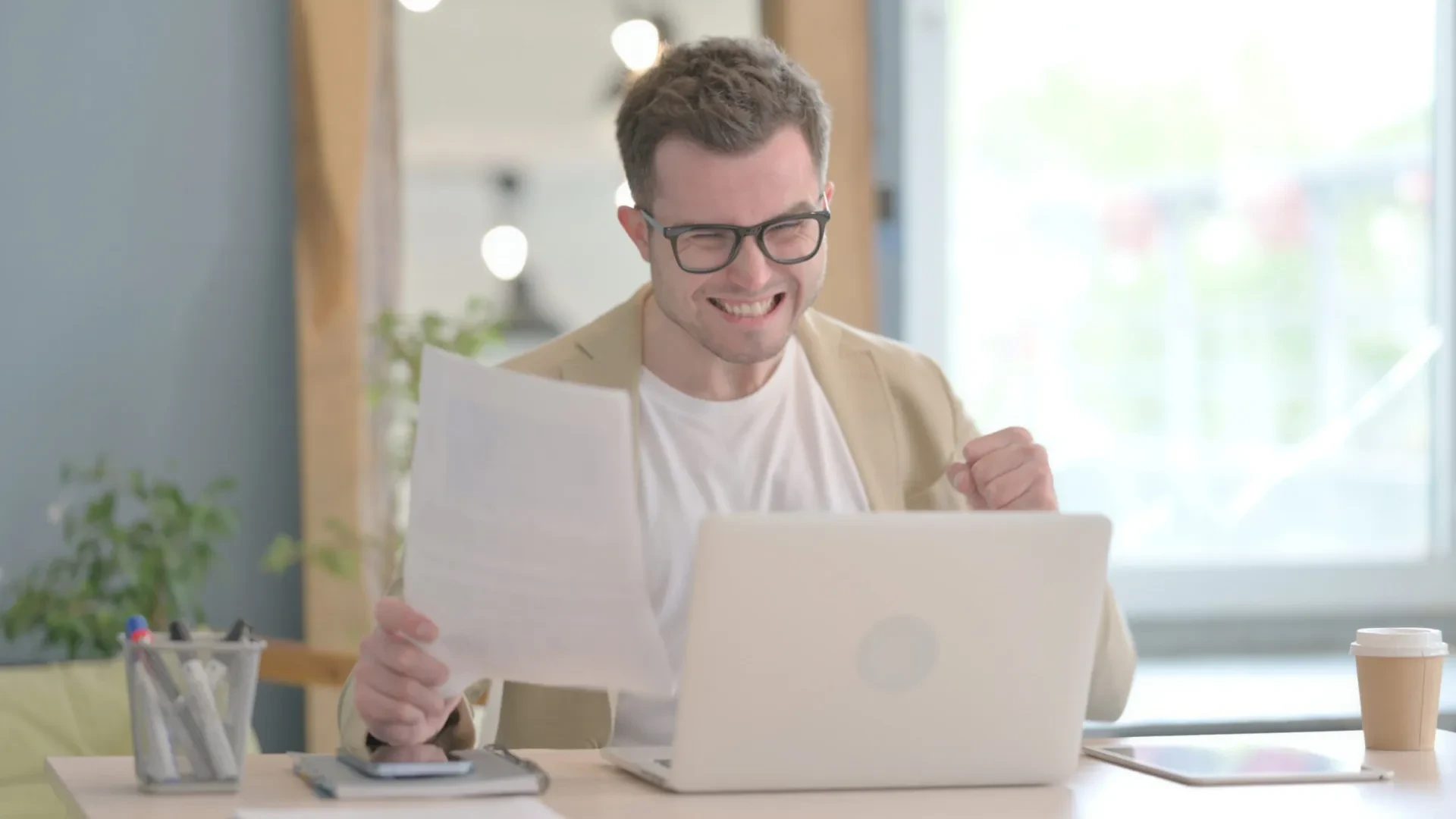 Lächelnder Mann mit Brille hält Dokument vor Laptop am Schreibtisch