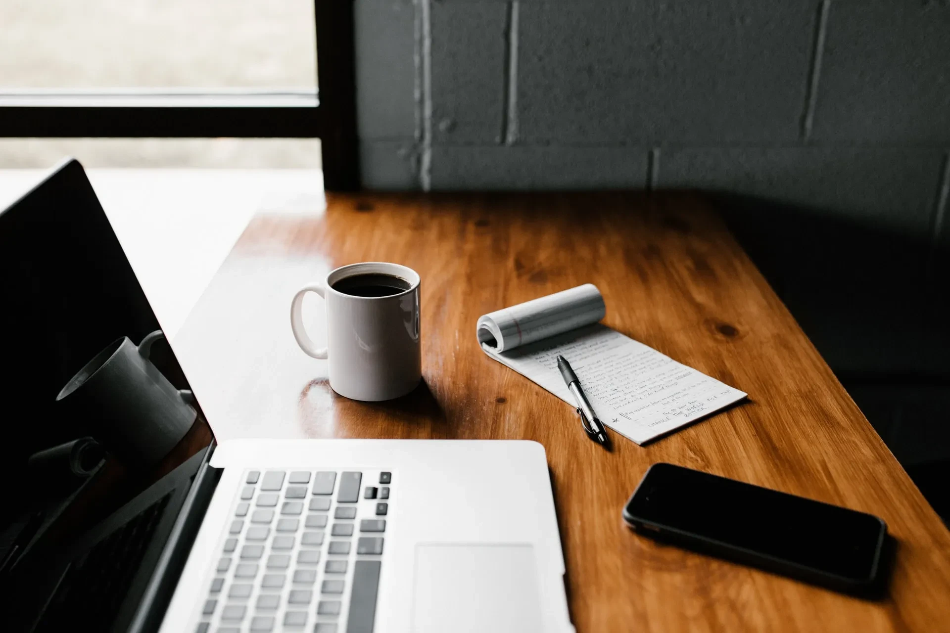 Laptop, Kaffeetasse, Notizbuch und Smartphone auf einem Holzschreibtisch
