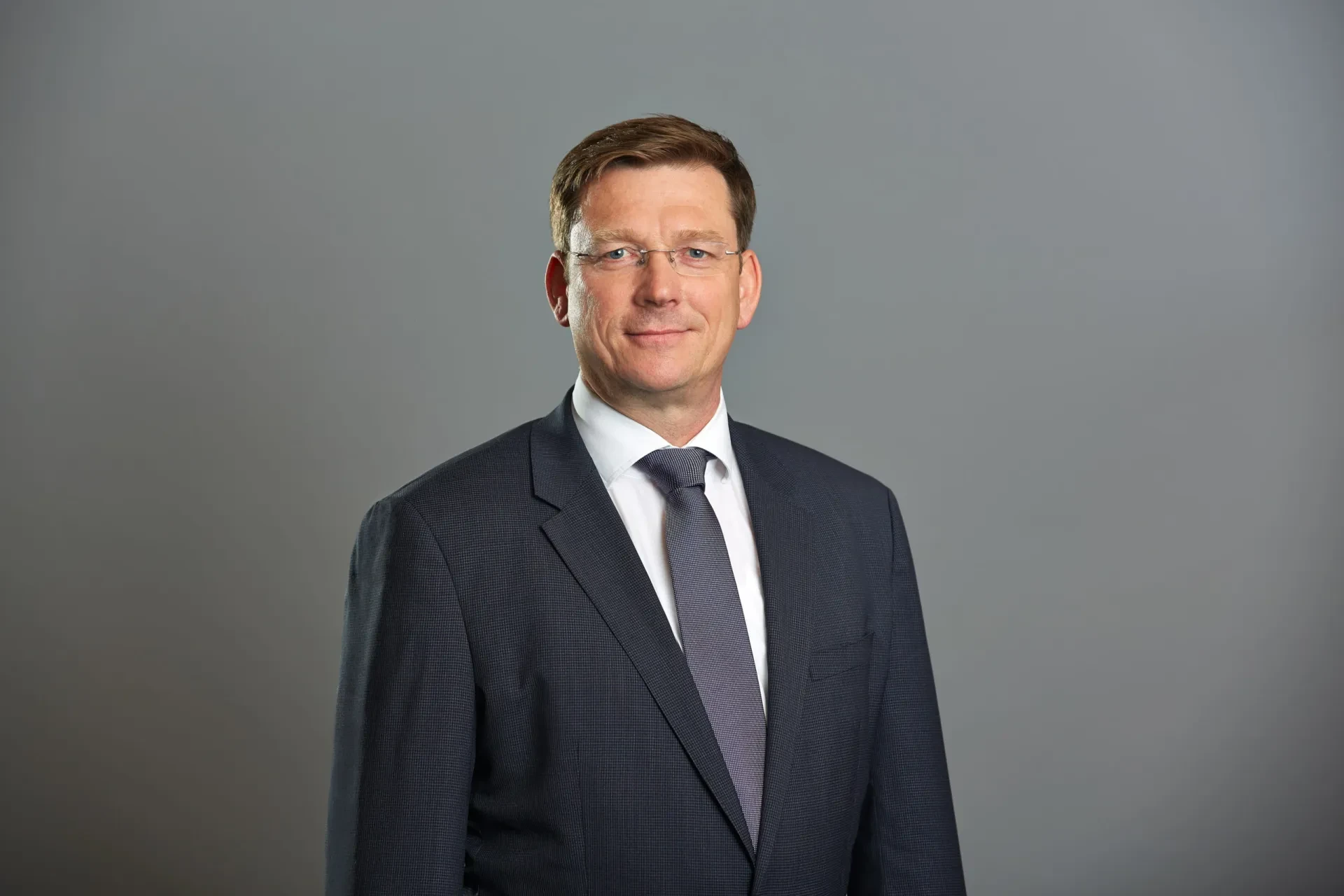 Ein Mann im grauen Anzug mit Brille, leichtes Lächeln.