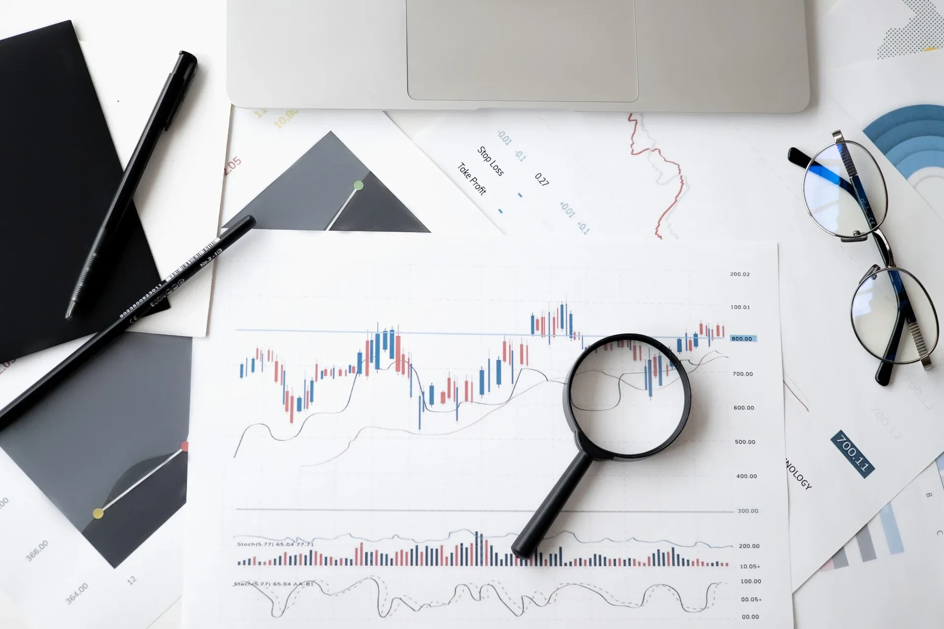 Finanzcharts mit Lupe, Brille und Laptop auf weißem Schreibtisch