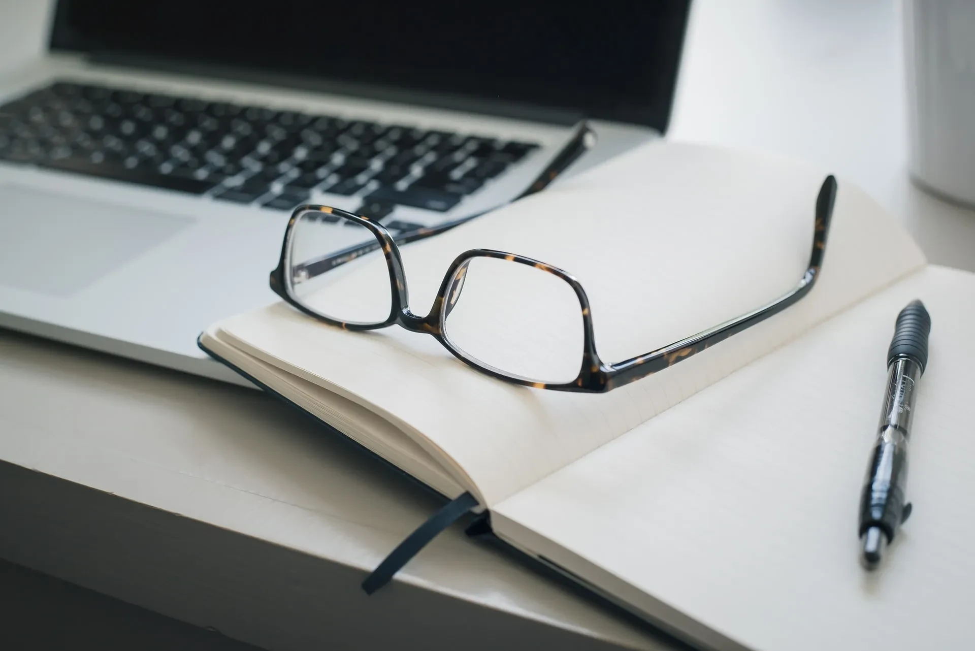 Brille auf aufgeschlagenem Notizbuch mit Stift und Laptop im Hintergrund