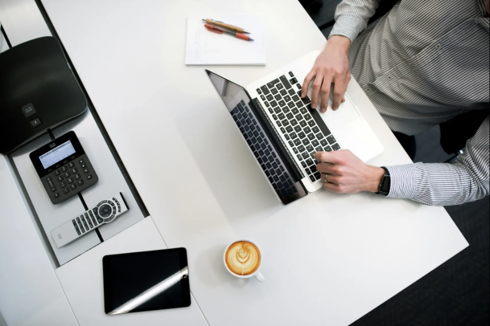 Vogelperspektive auf Person am Laptop mit Kaffee, Telefon und Notizbuch auf weißem Schreibtisch